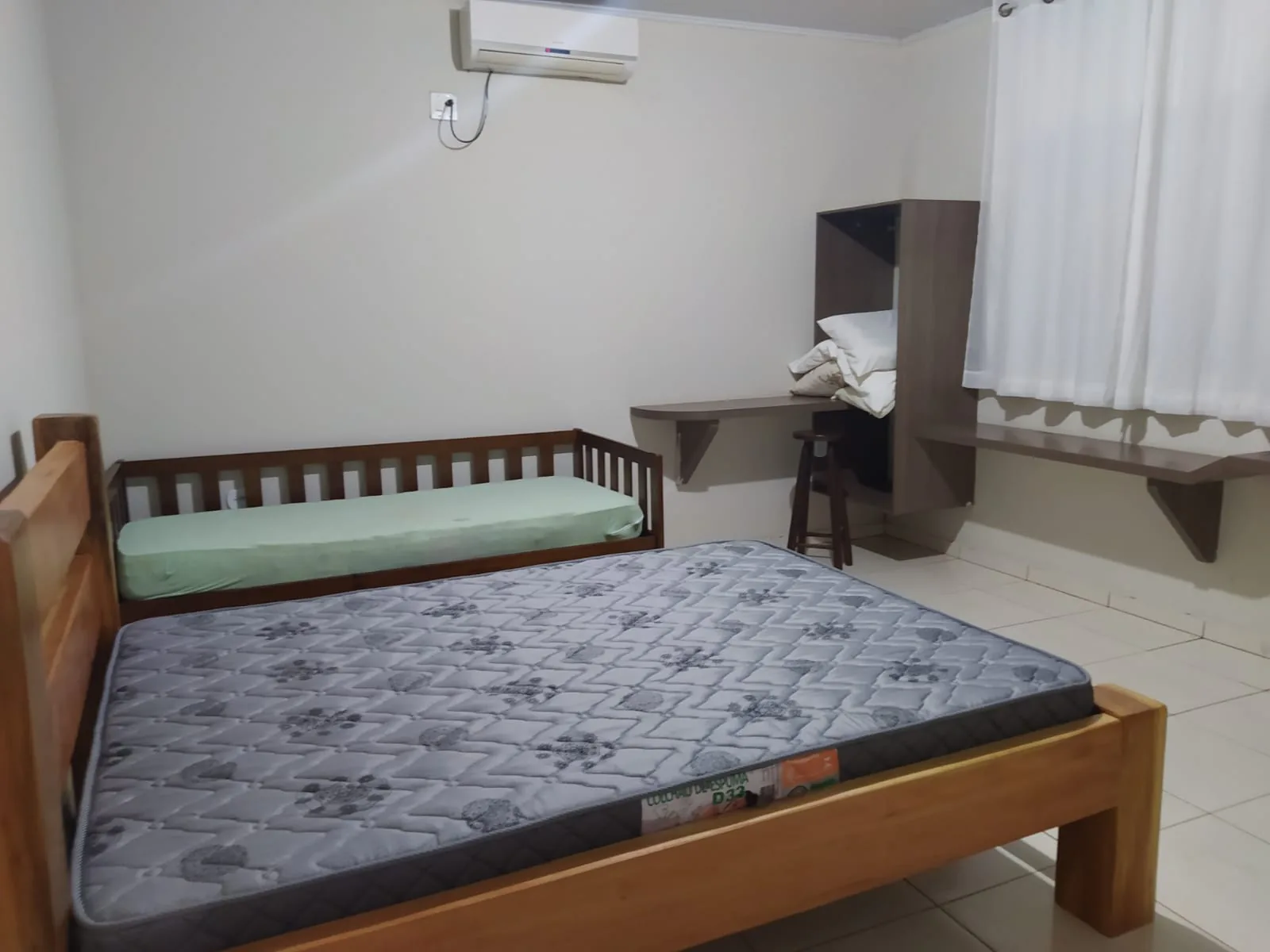 Interior da suíte: cama de casal, bancada com banqueta, nicho com roupa de cama e janela com cortina