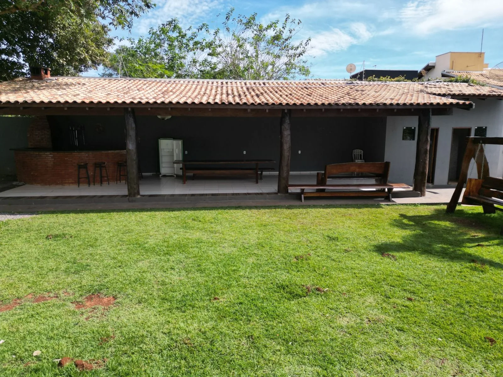 Varanda coberta do Espaço ToaToa vista do gramado: churrasqueira de tijolo, bar com banquetas, mesas de madeira e balanço